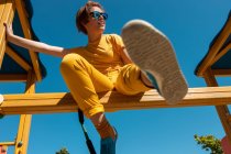 Знизу модний підліток в сонцезахисних окулярах сидить на жовтій поперечині на тлі чистого блакитного неба — стокове фото