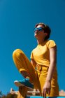Dal basso adolescente alla moda in occhiali da sole e abiti gialli eleganti seduti sulla traversa contro il cielo blu chiaro — Foto stock