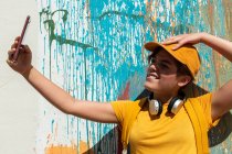 Stylish millennial taking selfie with smartphone while standing against wall with colorful paint drips — Stock Photo