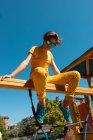 From below trendy teenager in sunglasses sitting on yellow crossbar against clear blue sky — Stock Photo