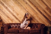 Junge Frau in weißem Kleid sitzt auf Couch mit Holzhintergrund. — Stockfoto