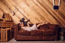 Молодая женщина записывает видео с винтажной камерой в интерьере с диваном на деревянном фоне — стоковое фото