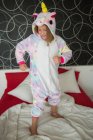 Happy joyful girl in funny kigurumi unicorn pajama playing in bedroom and jumping on bed with red and white bedding — Stock Photo