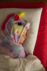 Lindo niño pequeño en colorido pijama unicornio kigurumi cubierto con manta durmiendo en la cama con ropa de cama blanca y roja - foto de stock