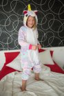 Menina alegre no pijama unicórnio se divertindo na cama com roupa de cama branca e vermelha — Fotografia de Stock