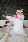 Fille gaie en pyjama licorne s'amuser et couvrir le visage sur le lit avec literie blanche et rouge — Photo de stock