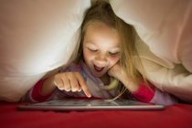 Muchacha preescolar emocionada usando tableta debajo de la manta en la cama - foto de stock