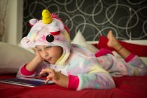 Chica en pijama unicornio usando tableta en la cama - foto de stock