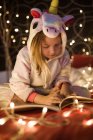 Маленькая симпатичная девочка читает книгу в спальне, украшенной рождественскими огнями — стоковое фото