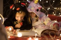 Graziosi fratelli in accogliente pigiama kigurumi leggere fiabe libro mentre seduti insieme sul letto decorato con luci di Natale — Foto stock