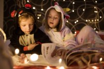 Irmãos bonitos em pijama kigurumi acolhedor ler contos de fadas livro enquanto sentados juntos na cama decorado com luzes de Natal — Fotografia de Stock