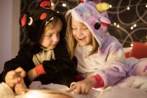 Lindos hermanos en acogedor pijama kigurumi leer cuentos de hadas libro mientras se sientan juntos en la cama decorada con luces de Navidad - foto de stock