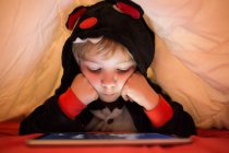 Petit garçon d'âge préscolaire en pyjama kigurumi utilisant une tablette sous la couverture au lit — Photo de stock