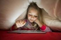 Linda niña rubia en pijama usando tableta mientras está acostada debajo de una manta en la cama por la noche - foto de stock