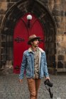Спокойный фотограф в шляпе прогуливается с камерой на булыжной улице против красной двери старого здания в Шотландии — стоковое фото
