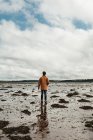 Обратный вид человека, идущего вдоль мокрого пляжа среди морских водорослей на фоне размытого горизонта и облачного неба в дневное время в Шотландии — стоковое фото