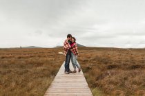 Coppia di viaggiatori in plaid in piedi su sentiero di legno tra palude marrone in Scozia su cupo giorno nuvoloso — Foto stock