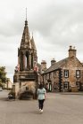 Vista posteriore di viaggiatore femminile a piedi sulla piazza della città vecchia con bella fontana medievale e edifici in pietra in Scozia — Foto stock
