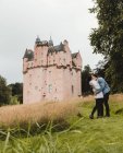Vista lateral de la feliz pareja abrazando de pie en verdes colinas junto al castillo medieval durante el viaje a Escocia - foto de stock