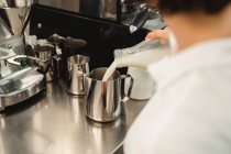 Cropped of female barista pouring milk from glass bottle to metal pot while working at counter with coffee equipment in cafe — Stock Photo