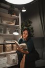 Joven mujer elegante sentada en silla en acogedora habitación cerca de la lámpara y libro de lectura con novela favorita mientras descansa en casa - foto de stock