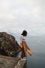 Боковой вид задумчивой одинокой женщины в повседневной одежде, балансирующей на краю скалы и смотрящей на размытый скалистый холм и спокойную океанскую воду в ветреную пасмурную погоду — стоковое фото