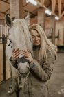 Длинноволосая женщина обнимает серую лошадь с белой гривой в морде и смотрит в камеру в стойле — стоковое фото