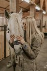 Длинноволосая женщина обнимает серую лошадь с белой гривой в конюшне — стоковое фото