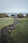 Сверху анонимная женщина наслаждается удивительным живописным ландшафтом Северной Ирландии во время путешествия, прогуливаясь возле быстрой мелководной реки, текущей к водам скалистого берега — стоковое фото