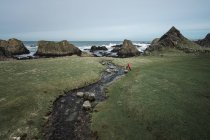 De cima mulher anônima desfrutando paisagem cênica incrível da Irlanda do Norte durante a viagem enquanto caminhava perto do rio raso rápido que flui para águas de costa rochosa — Fotografia de Stock