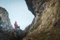 Rückansicht einer Frau in warmem Outfit, die auf einer Felskante in einer Höhle im Hafen von Nordirland sitzt und mit dem Handy ins Meer blickt — Stockfoto