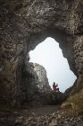 Vista trasera de la hembra en traje cálido sentada en el borde del acantilado dentro de la cueva en el puerto de Irlanda del Norte mirando hacia el mar utilizando el teléfono móvil - foto de stock