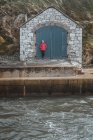Turista femenina en traje cálido apoyada en un antiguo edificio de piedra con puertas mientras está de pie en el pintoresco muelle del puerto de Ballintoy sobre fondo de rocas - foto de stock