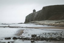 Paisagem cênica com Mussenden Temple localizado em penhasco de pedra na costa da Irlanda do Norte e ondas de mar tempestuosas colidindo contra rochas com céu cinza nublado no fundo — Fotografia de Stock