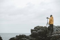 Rückansicht eines männlichen Reisenden, der mit Kamera auf Stativ auf einem Felsen steht und an bewölkten, düsteren Tagen an der nordirischen Küste das Meer fotografiert — Stockfoto