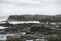 Мужчина-путешественник, стоящий на скале с камерой на штативе и фотографирующий морской пейзаж в пасмурный день на побережье Северной Ирландии — стоковое фото
