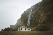 Kleines ländliches Holzhaus mit weißen Wänden und gelbem Satteldach auf einer grünen Wiese am Fuße der Klippe mit Wasserfall vor grauem wolkenverhangenem Himmel an einem Frühlingstag in Nordirland — Stockfoto
