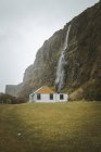 Small country wooden house with white walls and yellow gabled roof located on green meadow at foot of cliff with waterfall against grey cloudy sky in spring day in Northern Ireland — Stock Photo
