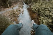 Desde arriba viajero de cultivos en jeans y zapatillas de deporte sentado en el borde del puente y las piernas colgando sobre el río durante el senderismo en el parque forestal de primavera en Irlanda del Norte - foto de stock
