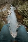 From above crop traveler in jeans and sneakers sitting on edge of bridge and dangling legs over river during hiking in spring forest park in Northern Ireland — Stock Photo