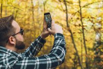 Uomo in camicia scozzese scattare foto di alberi autunnali sul telefono cellulare con boschi su sfondo sfocato — Foto stock