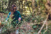 Longo cabelo feminino hipster em elegante casual xadrez camisa tênis e óculos de sol na moda sentado em pequeno toco em spangled de caminho de folhagem entre árvores verdes — Fotografia de Stock