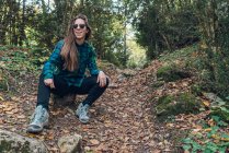 Long haired female hipster in stylish casual plaid shirt sneakers and trendy sunglasses sitting on small stub on spangled of leafage path among green trees — Stock Photo