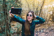 Mulher de camisa xadrez tirando selfie no celular enquanto está de pé na floresta de madeira — Fotografia de Stock