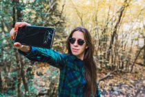 Woman in plaid shirt taking selfie on mobile phone while standing on forest wood — Stock Photo