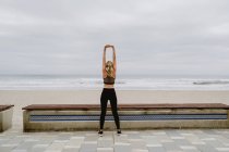Rückansicht einer Sportlerin in aktiver Kleidung, die bei bewölktem Wetter mit erhobenen Händen in Küstennähe steht — Stockfoto