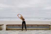 Вид сзади спортсменки в активной одежде, стоящей с поднятыми руками и растянувшейся на берегу моря в пасмурную погоду — стоковое фото