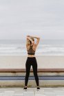 Vista posteriore dell'atleta donna in abbigliamento attivo in piedi con le mani sollevate e che si estende vicino alla riva del mare su tempo nuvoloso — Foto stock