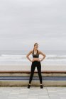 Женщина-атлет в активной одежде стоит с руками на бедрах возле побережья в пасмурную погоду — стоковое фото