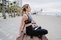 Vista lateral de atleta senhora concentrada sentado no banco e olhando para longe com o litoral no fundo borrado — Fotografia de Stock